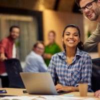 Two young multiracial colleagues looking at camera and smiling while working together on laptop in the modern office or coworking space. Teamwork, cooperation, business concept